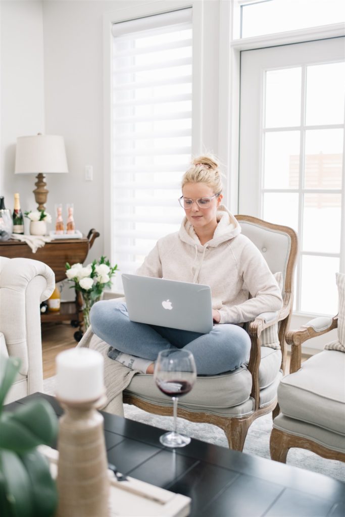 A woman sits on her laptop in a living room with glasses on