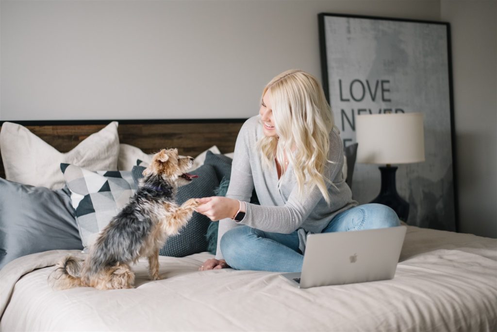 Blonde woman sitting on bed with laptop holding a small dogs paw during monthly branding session