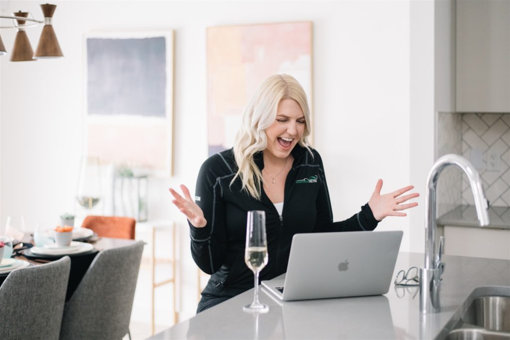 Blonde woman exclaiming happily while looking at laptop with a glass of champagne during monthly branding session