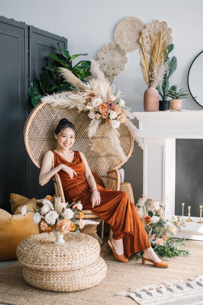 woman surrounded by flowers sitting in a wicker chair wearing a burnt orange gown