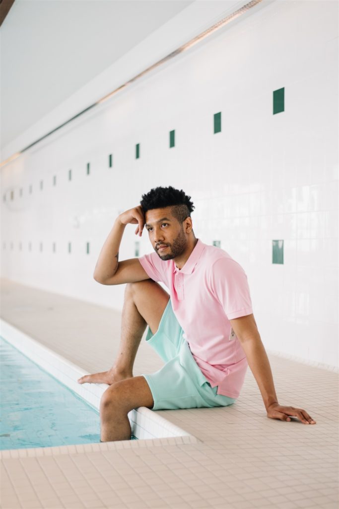 Man wearing baby blue swim shorts and a pink polo sitting with his legs in an indoor pool