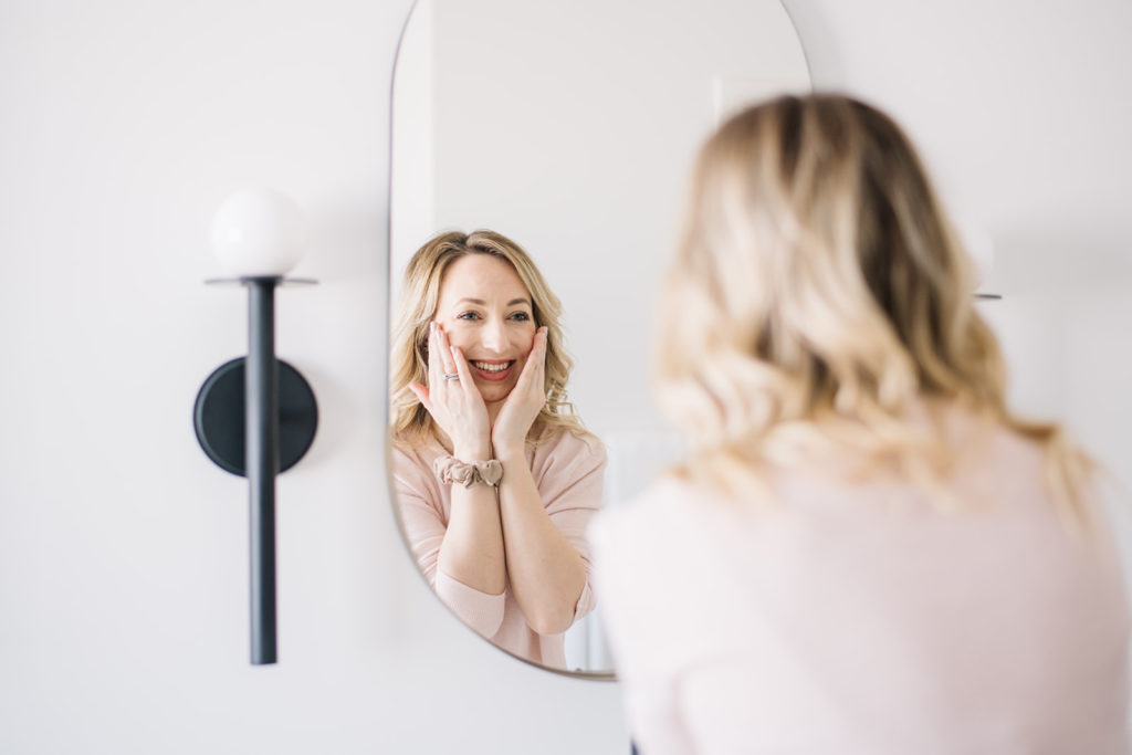 woman smiling into a mirror as she applies a skincare product to her cheeks