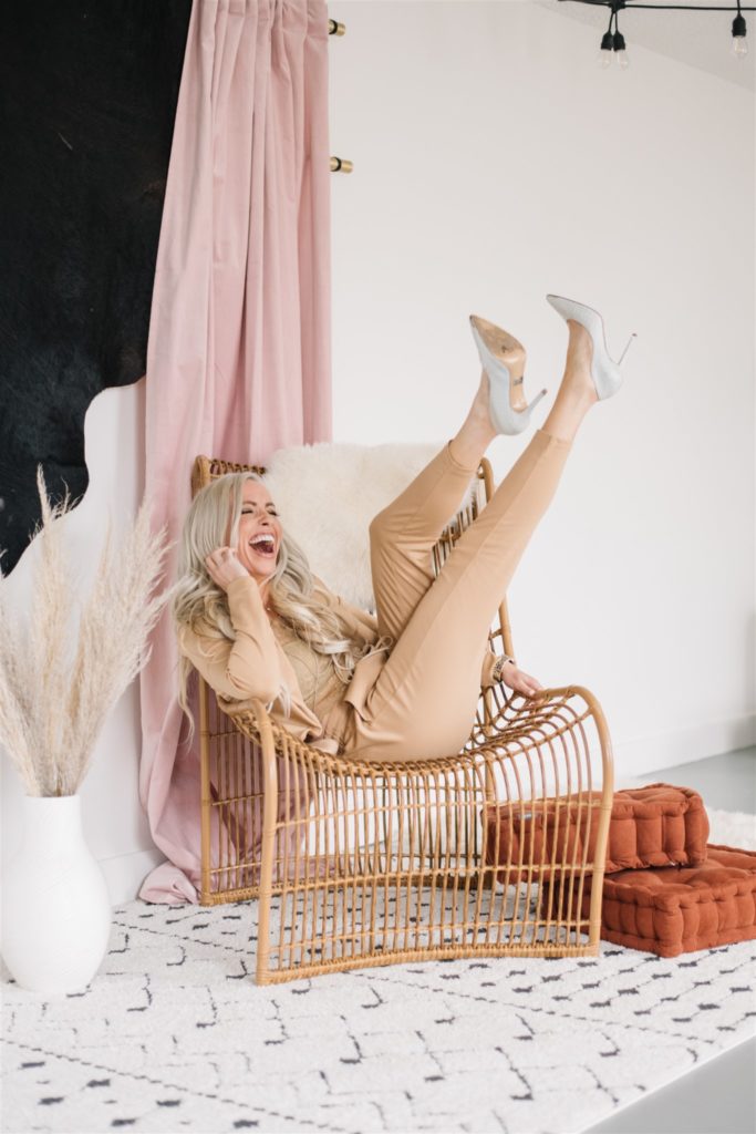 Blonde woman laughing on the phone with her feet in the air as she sits in a wicker chair