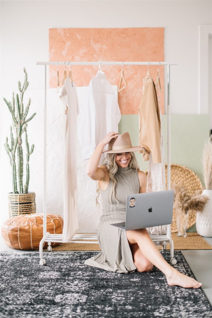 Woman sitting on a rug holding her hat on her head with one hand and her laptop in her other hand