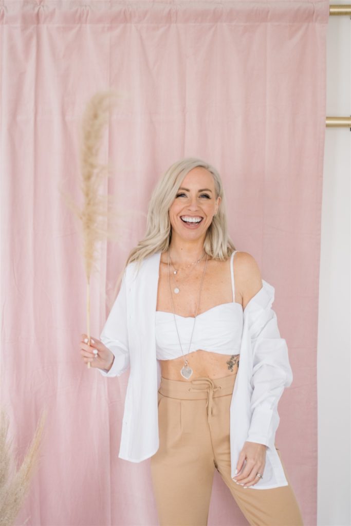 Blonde woman laughing on front of a pink backdrop holding a piece of pampas grass