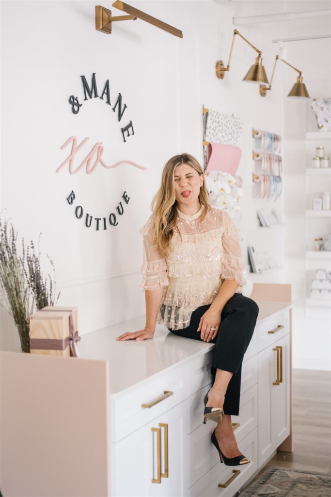 Woman sticks out her tongue as she sits on the counter next to XO & Mane Boutique sign